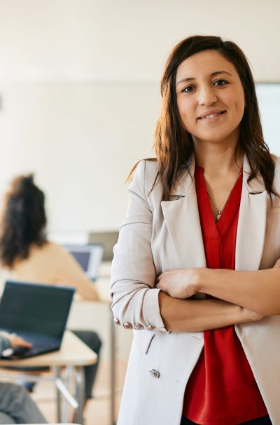 Happy female professor standing with arms crossed during computer class and looking at camera. Her students are e-learning in the background.